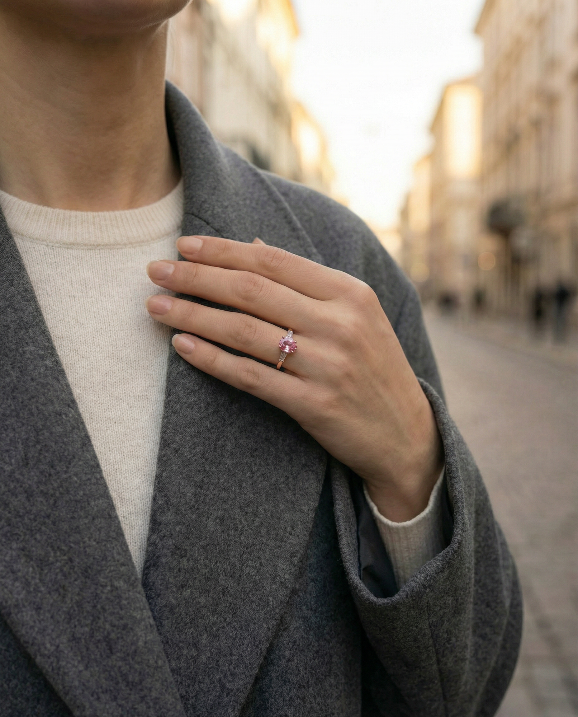 Woman wearing a grey coat with a blurred street scene in the background. Wearing a pink sapphire and diamond engagement ring on her ring finger.
